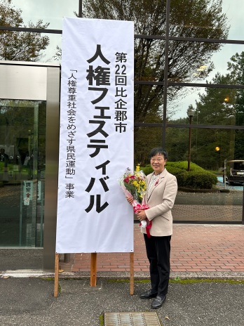 人権フェスティバルの看板の前で花束を持っている萩原なつ子の写真